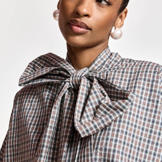 A close-up of a model wearing a brown, beige, and white checkered blouse with a large bow tie at the collar, paired with white pearl earrings.