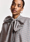 A close-up of a model wearing a brown, beige, and white checkered blouse with a large bow tie at the collar, paired with white pearl earrings.