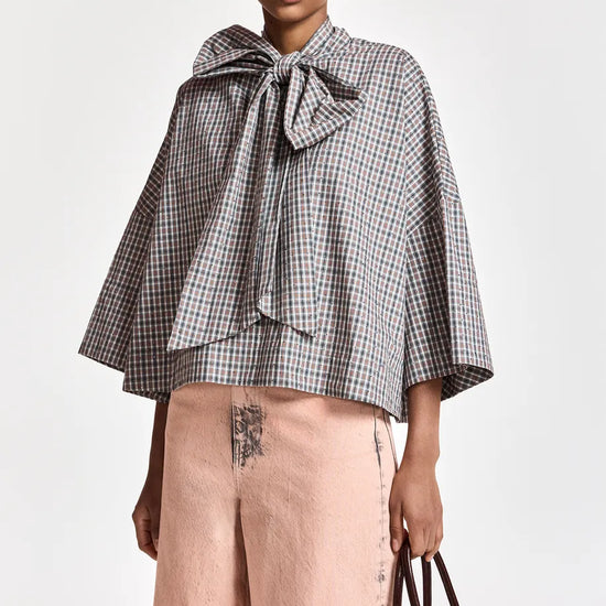 A model wearing a black and white gingham cape-style blouse with a large bow at the collar, paired with dusty pink wide-leg trousers and holding a dark brown handbag against a plain light background.