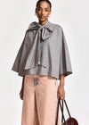 A model wearing a black and white gingham cape-style blouse with a large bow at the collar, paired with dusty pink wide-leg trousers and holding a dark brown handbag against a plain light background.