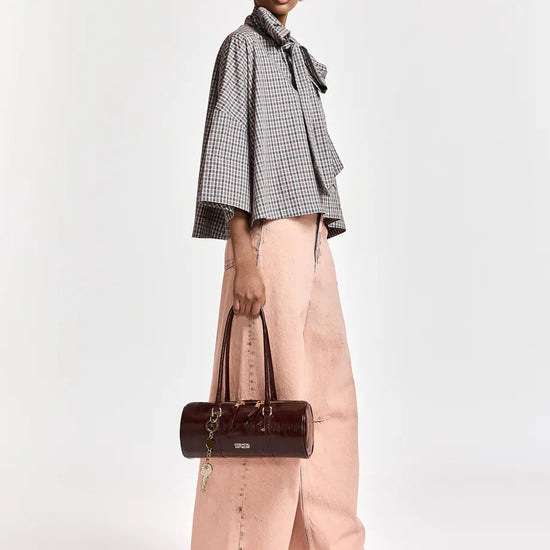 A model is shown from the side wearing a black and white checkered bow-tie blouse, wide-leg dusty pink trousers, and holding a dark brown leather cylindrical handbag against a plain white studio background.