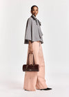 A model is shown from the side wearing a black and white checkered bow-tie blouse, wide-leg dusty pink trousers, and holding a dark brown leather cylindrical handbag against a plain white studio background.