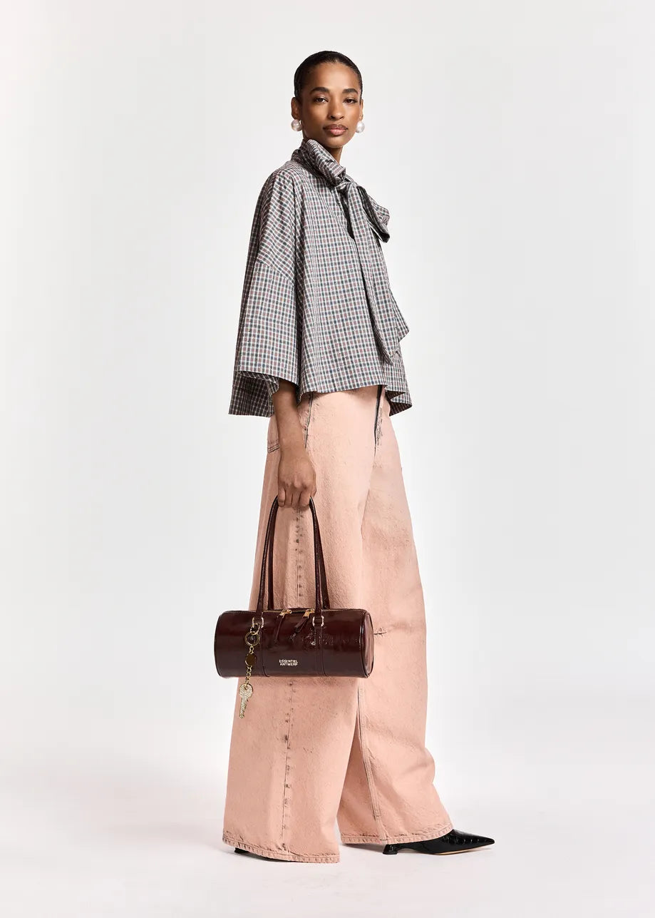 A model is shown from the side wearing a black and white checkered bow-tie blouse, wide-leg dusty pink trousers, and holding a dark brown leather cylindrical handbag against a plain white studio background.
