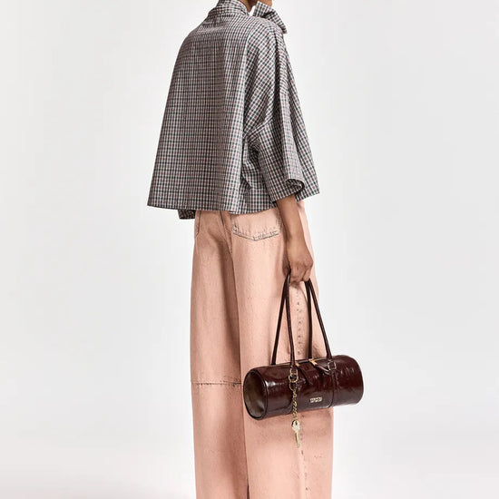 A model is shown from a side-back angle wearing a black and white checkered cape jacket, light pink wide-leg pants, and holding a dark brown cylindrical handbag against a plain light gray background.