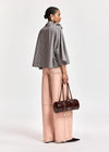 A model is shown from a side-back angle wearing a black and white checkered cape jacket, light pink wide-leg pants, and holding a dark brown cylindrical handbag against a plain light gray background.