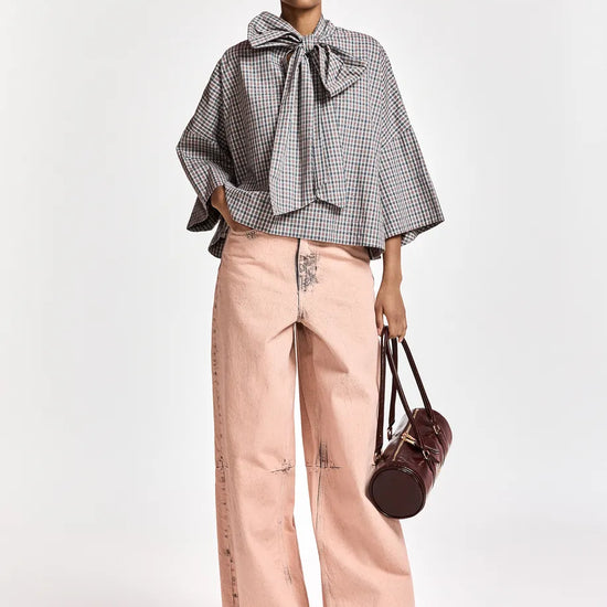 A model stands against a light gray studio background wearing a gray and white gingham cape-style blouse with a large bow at the collar, light pink wide-leg pants, black pointed shoes, and holding a dark brown handbag.