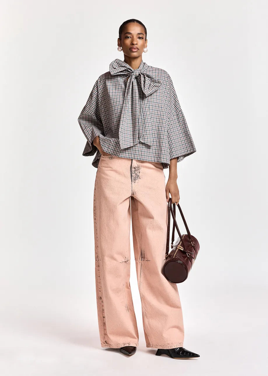 A model stands against a light gray studio background wearing a gray and white gingham cape-style blouse with a large bow at the collar, light pink wide-leg pants, black pointed shoes, and holding a dark brown handbag.