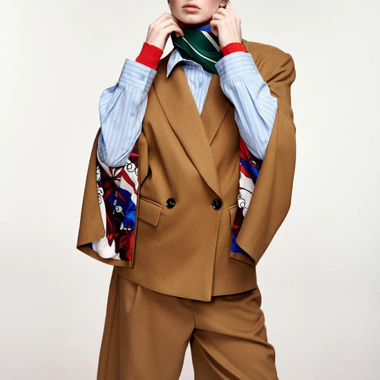 A model wearing a brown double-breasted blazer and matching cropped trousers, layered over a light blue striped shirt, a green and red striped tie, and a colorful patterned scarf, against a plain light gray background.