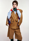 A model wearing a brown double-breasted blazer and matching cropped trousers, layered over a light blue striped shirt, a green and red striped tie, and a colorful patterned scarf, against a plain light gray background.
