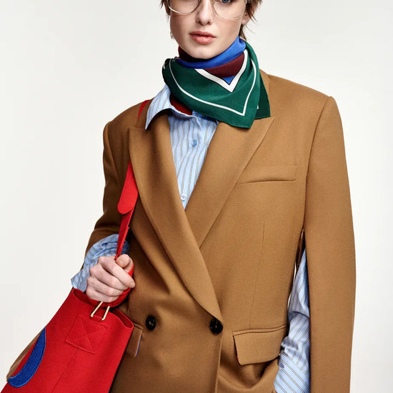 A model wearing a camel brown double-breasted blazer, a light blue striped shirt, a green and white patterned neck scarf, and holding a bright red tote bag with blue accents, set against a plain light gray background.