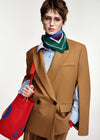 A model wearing a camel brown double-breasted blazer, a light blue striped shirt, a green and white patterned neck scarf, and holding a bright red tote bag with blue accents, set against a plain light gray background.