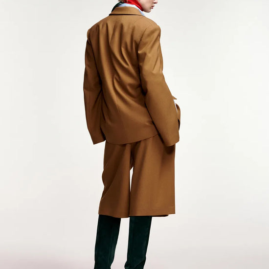 A model shown from the back wearing a camel brown tailored suit with a matching blazer and trousers, paired with dark green over-the-knee boots and a red neck scarf, against a plain white studio background.