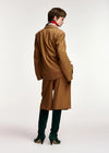 A model shown from the back wearing a camel brown tailored suit with a matching blazer and trousers, paired with dark green over-the-knee boots and a red neck scarf, against a plain white studio background.
