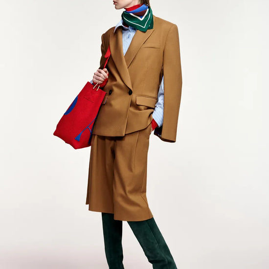 A model is shown in profile wearing a camel brown tailored suit with a matching knee-length skirt, a green striped scarf, a light blue shirt, dark green velvet knee-high boots, and holding a red and blue striped tote bag against a plain light gray background.