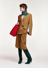 A model is shown in profile wearing a camel brown tailored suit with a matching knee-length skirt, a green striped scarf, a light blue shirt, dark green velvet knee-high boots, and holding a red and blue striped tote bag against a plain light gray background.