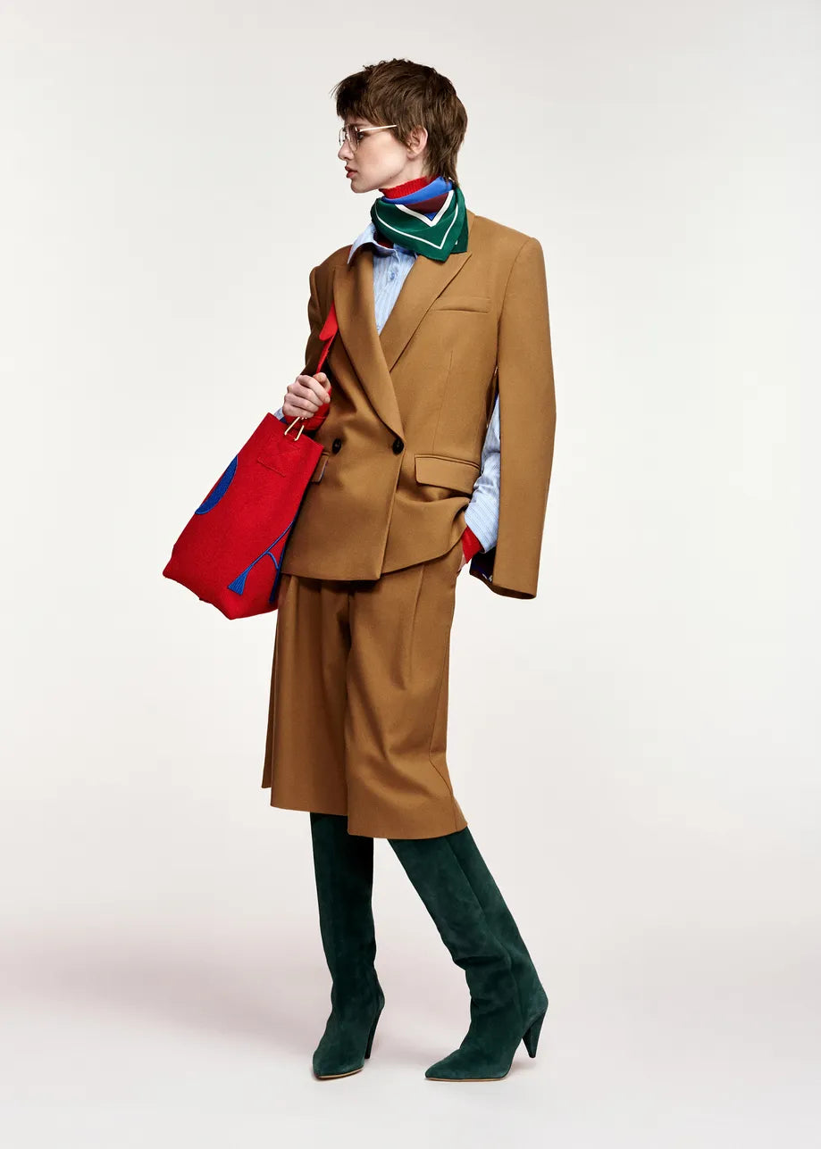 A model is shown in profile wearing a camel brown tailored suit with a matching knee-length skirt, a green striped scarf, a light blue shirt, dark green velvet knee-high boots, and holding a red and blue striped tote bag against a plain light gray background.