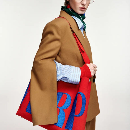A model wearing a camel brown blazer, striped shirt, and green scarf carries a large red tote bag with bold blue embroidered letters 'RP' on a plain light background.