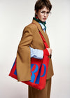 A model wearing a camel brown blazer, striped shirt, and green scarf carries a large red tote bag with bold blue embroidered letters 'RP' on a plain light background.