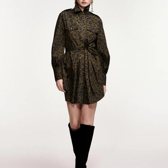 A model wearing a dark olive green and black leopard-print mini dress with a high collar and long sleeves, paired with black over-the-knee boots, shot against a plain light gray studio background.