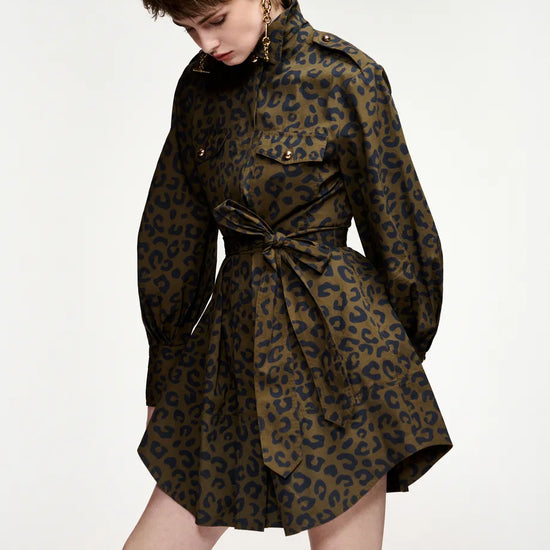 A model wearing a dark olive green and black leopard print mini dress with a tie waist belt, styled with black knee-high socks against a plain white studio background.