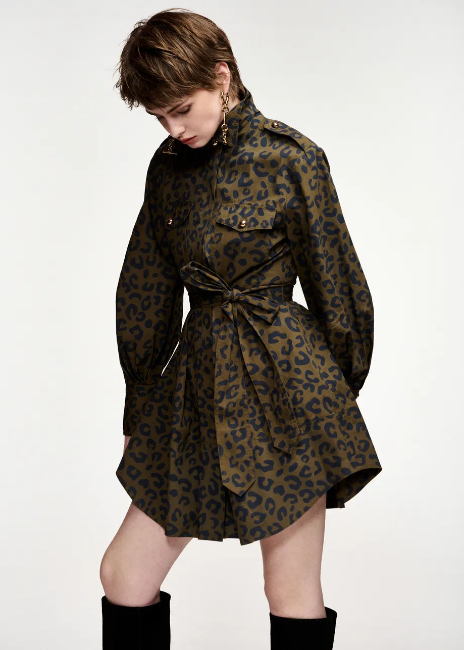A model wearing a dark olive green and black leopard print mini dress with a tie waist belt, styled with black knee-high socks against a plain white studio background.
