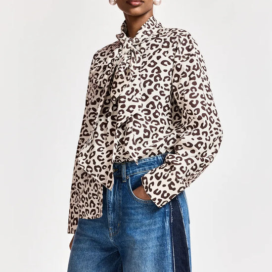 A model wearing a long-sleeve leopard print blouse with a ruffled high collar, paired with blue jeans, against a plain light gray background.