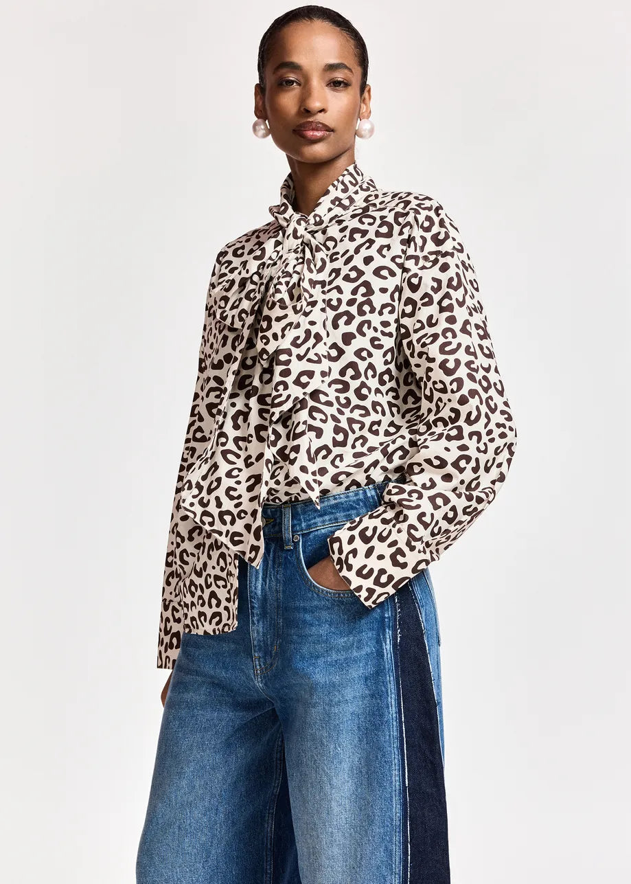 A model wearing a long-sleeve leopard print blouse with a ruffled high collar, paired with blue jeans, against a plain light gray background.