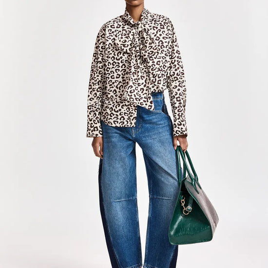A model stands against a light gray studio background wearing a beige and black leopard-print long-sleeve blouse with a tie-neck detail, wide-leg blue jeans, burgundy pointed-toe heeled boots, and holding a green tote bag.
