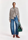 A model stands against a light gray studio background wearing a beige and black leopard-print long-sleeve blouse with a tie-neck detail, wide-leg blue jeans, burgundy pointed-toe heeled boots, and holding a green tote bag.