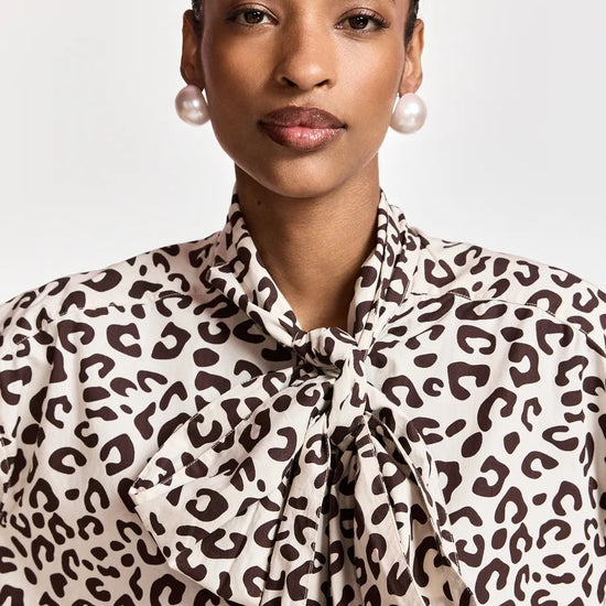 A close-up of a model wearing a leopard print blouse with a tie-neck detail, paired with pearl earrings, against a light gray background.