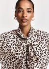 A close-up of a model wearing a leopard print blouse with a tie-neck detail, paired with pearl earrings, against a light gray background.