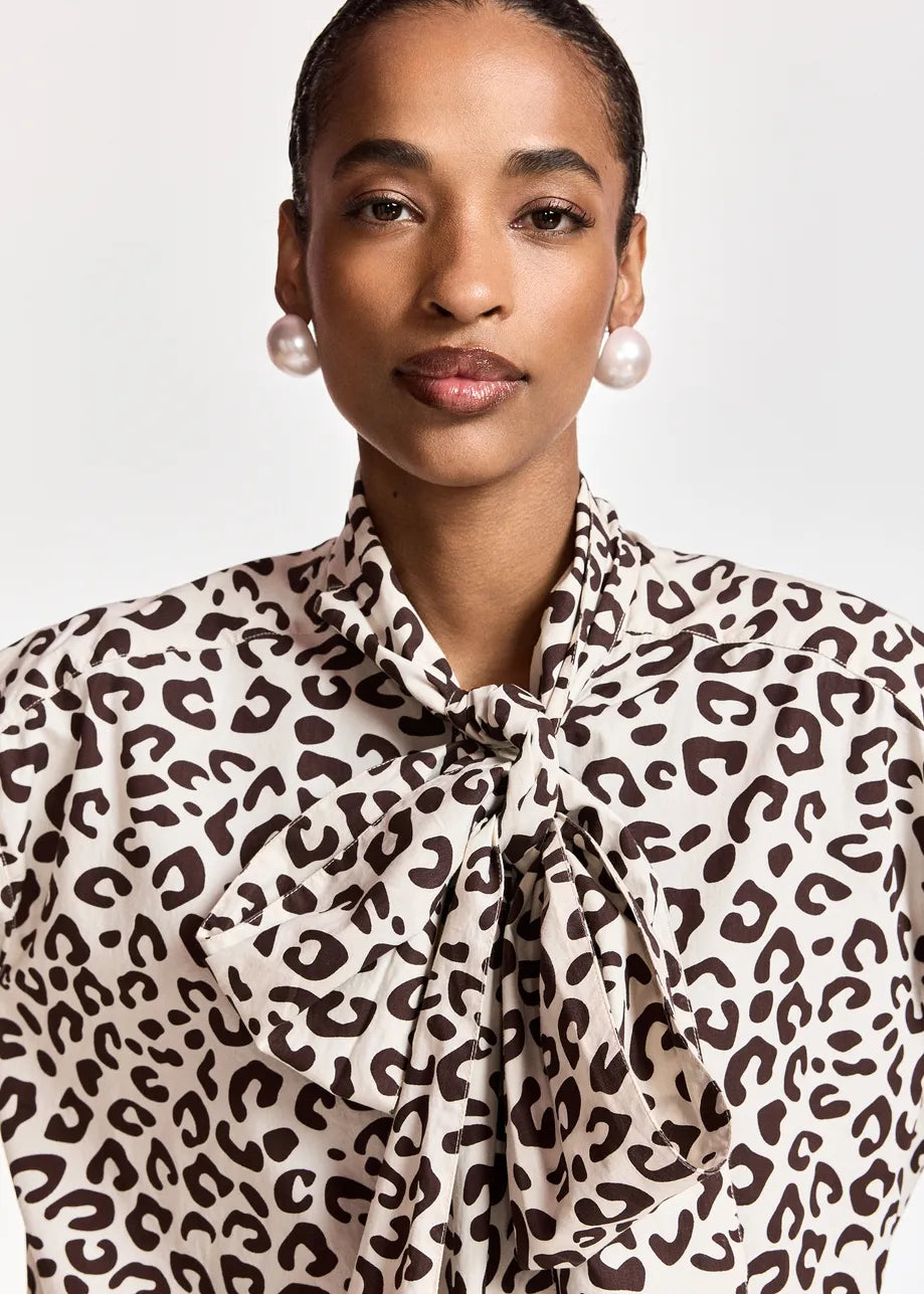 A close-up of a model wearing a leopard print blouse with a tie-neck detail, paired with pearl earrings, against a light gray background.