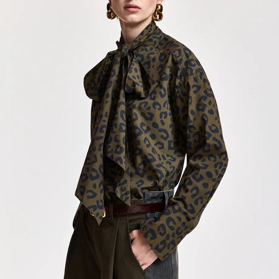 A model wearing a long-sleeve leopard print blouse with a large bow tie-neck collar, styled with dark wide-leg trousers and a dark belt against a plain light gray background.