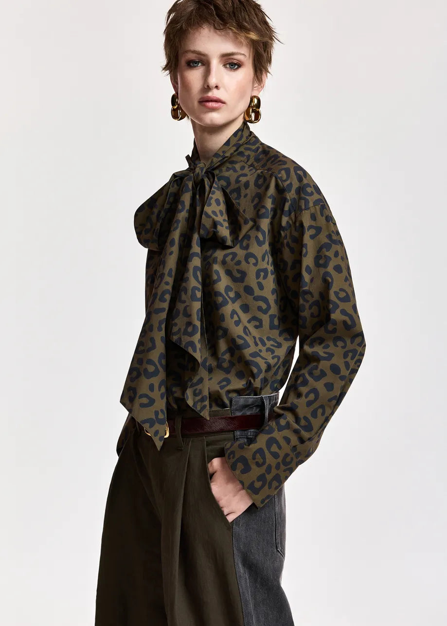 A model wearing a long-sleeve leopard print blouse with a large bow tie-neck collar, styled with dark wide-leg trousers and a dark belt against a plain light gray background.