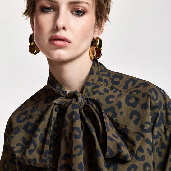 A close-up of a model wearing a leopard print blouse with a large bow tie-neck collar in olive green and navy blue, paired with chunky gold hoop earrings.