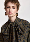 A close-up of a model wearing a leopard print blouse with a large bow tie-neck collar in olive green and navy blue, paired with chunky gold hoop earrings.