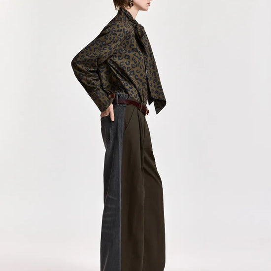 A model is shown in profile wearing a dark brown and black patterned long-sleeve shirt, dark wide-leg trousers, and dark pointed shoes against a plain light gray studio background.