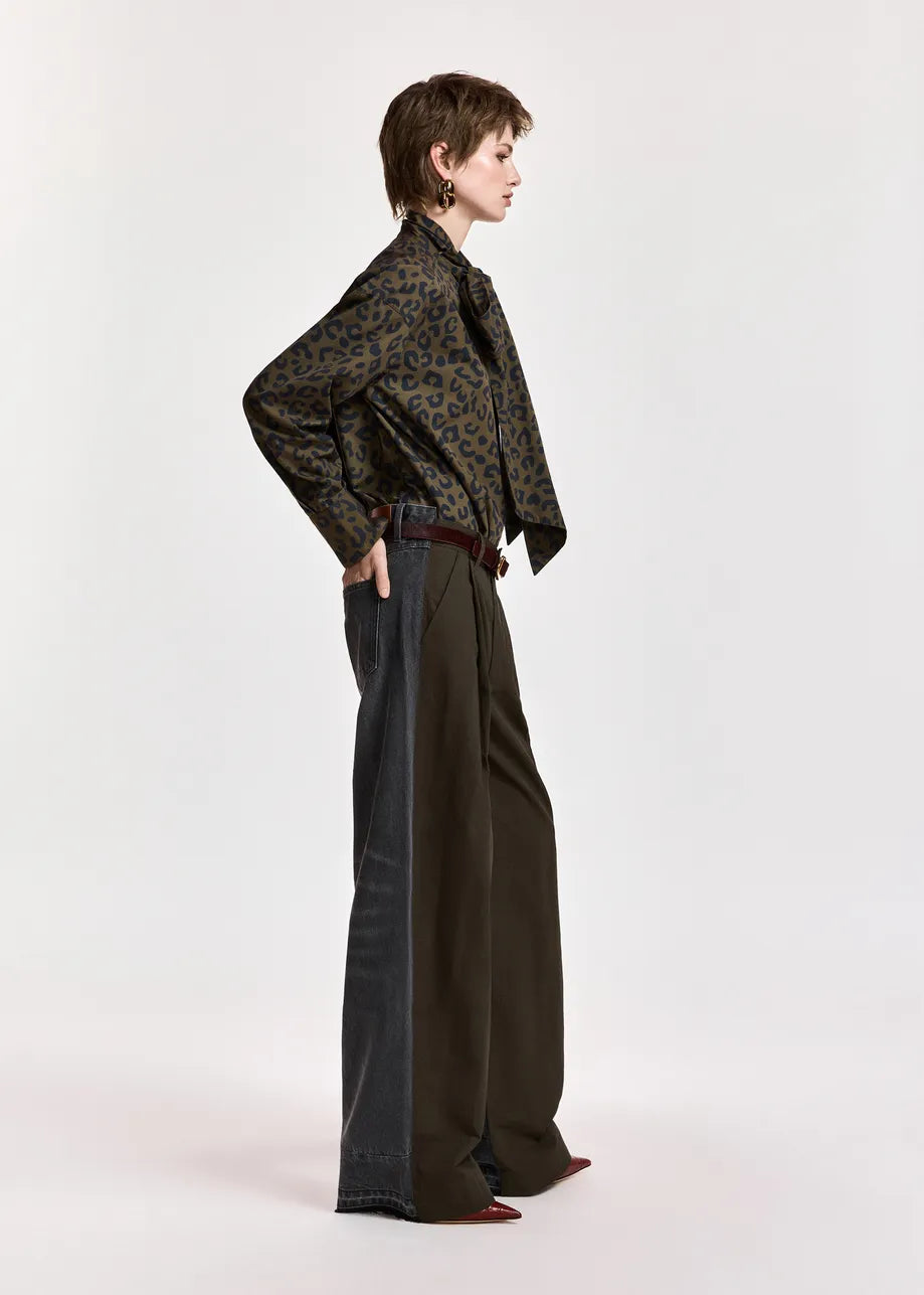 A model is shown in profile wearing a dark brown and black patterned long-sleeve shirt, dark wide-leg trousers, and dark pointed shoes against a plain light gray studio background.