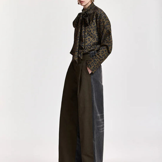 A model stands in profile wearing a dark patterned long-sleeve blouse with a high ruffled collar and wide-leg dark trousers against a plain light gray studio background.