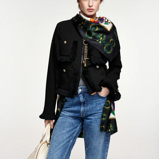 A model wearing a black double-breasted jacket with fringe trim, a colorful patterned scarf draped over the shoulder, blue jeans, and holding a cream-colored handbag against a light gray studio background.