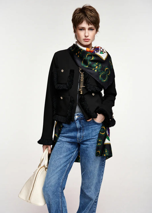A model wearing a black double-breasted jacket with fringe trim, a colorful patterned scarf draped over the shoulder, blue jeans, and holding a cream-colored handbag against a light gray studio background.