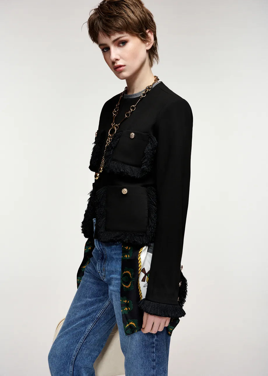 A model wearing a black fringed jacket with gold-tone hardware, layered over a gray top and paired with blue jeans, photographed against a plain light gray studio background.