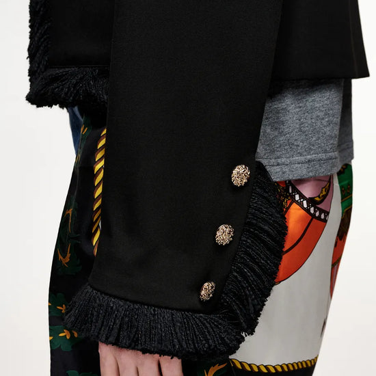 Close-up of a black fringed jacket or coat with ornate gold-tone decorative buttons on the sleeve, worn over a gray shirt and a colorful patterned garment, with blue jeans visible at the bottom.