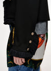 Close-up of a black fringed jacket or coat with ornate gold-tone decorative buttons on the sleeve, worn over a gray shirt and a colorful patterned garment, with blue jeans visible at the bottom.