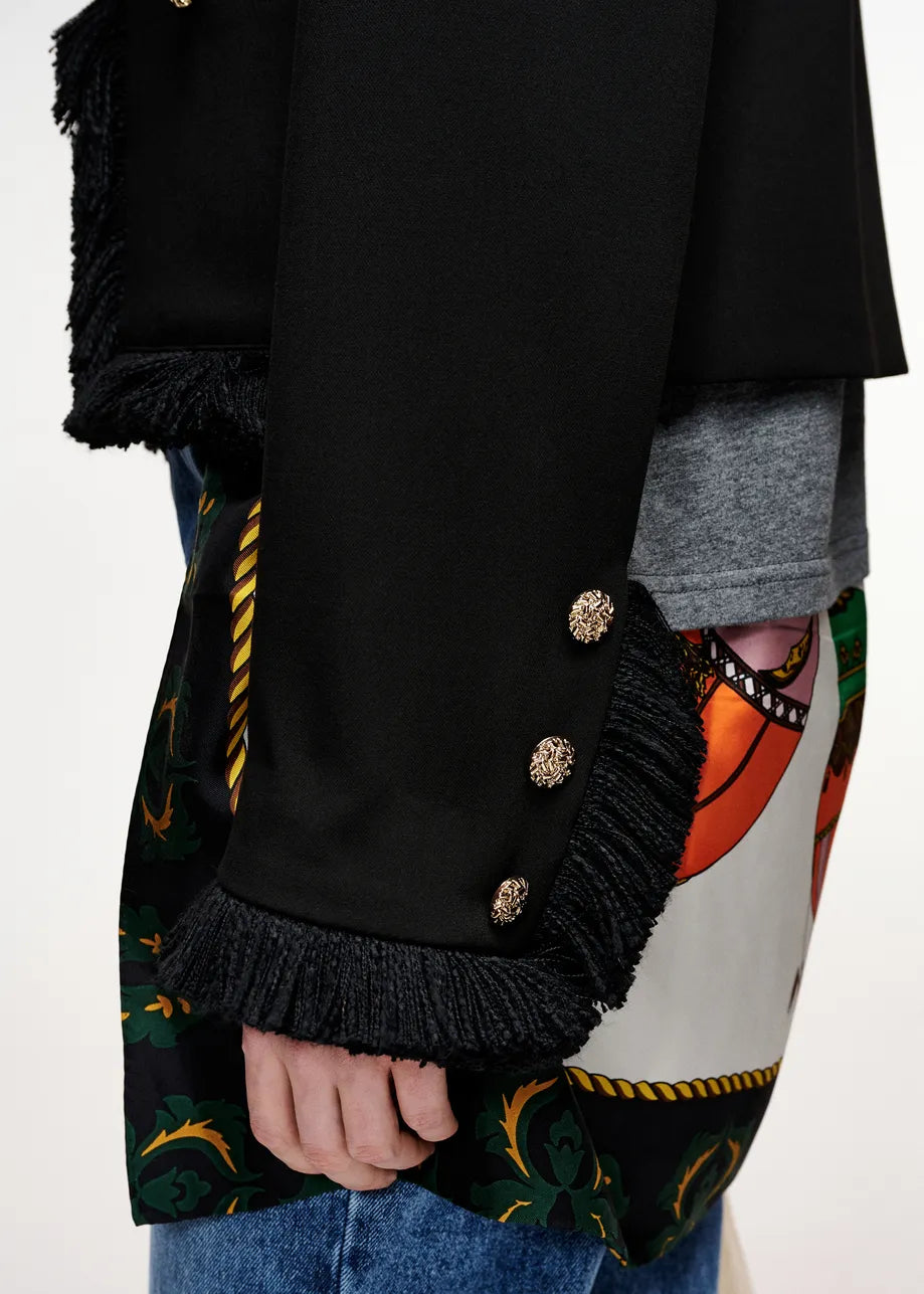 Close-up of a black fringed jacket or coat with ornate gold-tone decorative buttons on the sleeve, worn over a gray shirt and a colorful patterned garment, with blue jeans visible at the bottom.