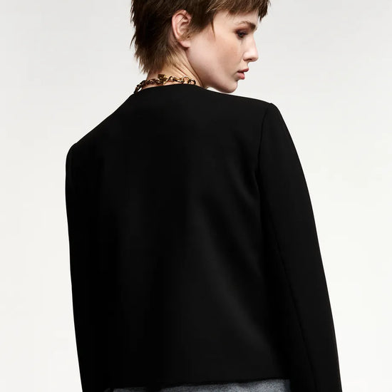 A model shown from the back wearing a black cropped jacket with long sleeves, layered over a gray top and a colorful patterned bottom, set against a plain light gray studio background.