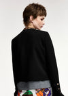 A model shown from the back wearing a black cropped jacket with long sleeves, layered over a gray top and a colorful patterned bottom, set against a plain light gray studio background.