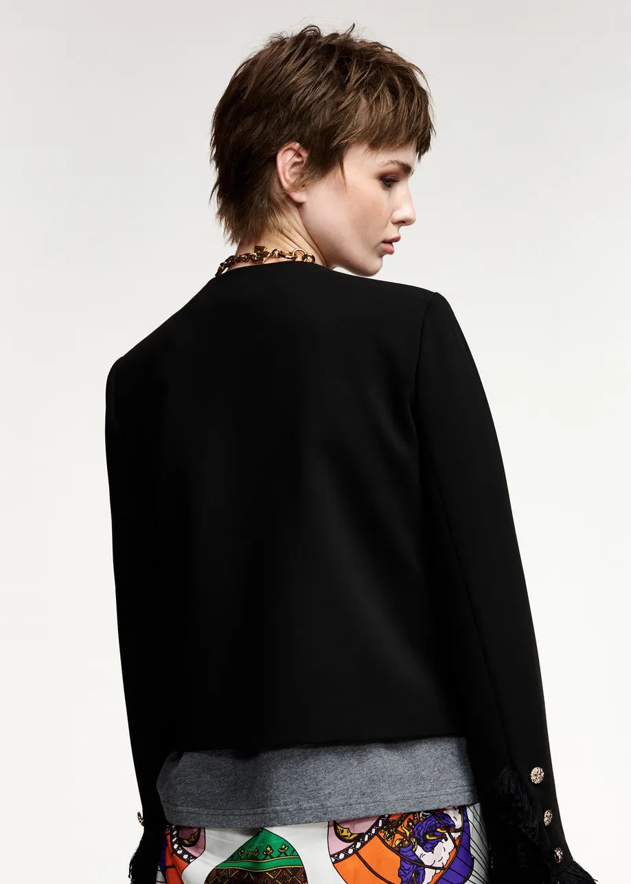 A model shown from the back wearing a black cropped jacket with long sleeves, layered over a gray top and a colorful patterned bottom, set against a plain light gray studio background.