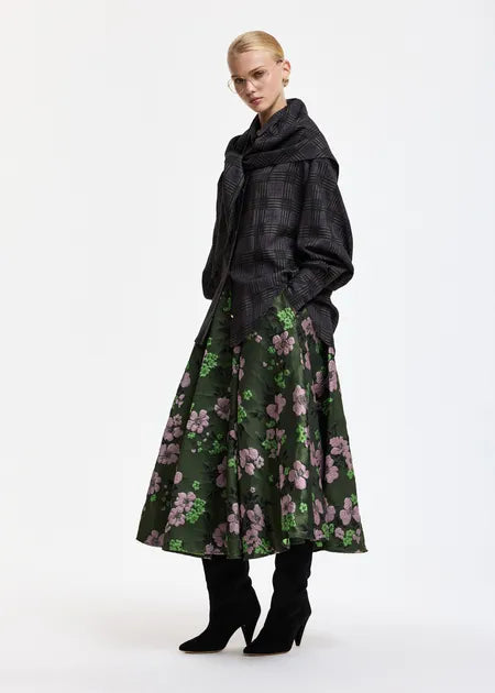 A model is shown from the back wearing a dark green midi skirt with a pink and green floral pattern, paired with a dark gray plaid shawl and black boots, against a plain white studio background.
