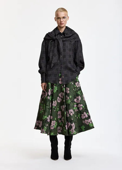 A model stands against a plain white studio background wearing a dark gray plaid shawl over a dark shirt and a dark green midi skirt with a pink floral print, paired with black boots.